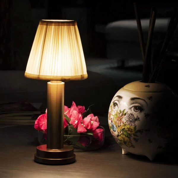 Lampe de chevet à l'abat-jour en tissu allumée, mise en scène avec des fleurs et un objet décoratif derrière, sur une table de chevet.