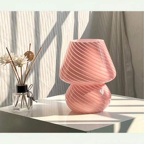 Lampe de chevet en verre soufflé, dans sa variante rose, sur une table de chevet et près d'une fenêtre.