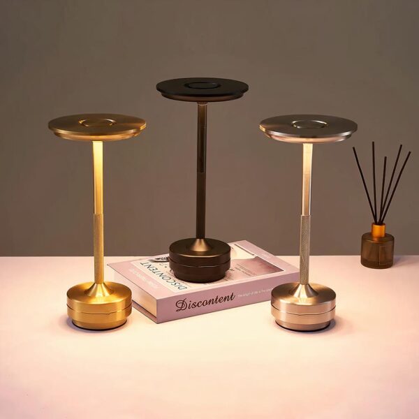 3 lampes design noir, doré et argenté posées sur une table blanche dont une sur un livre. Avec une décoration à côté.