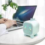 Lampe réveil dinosaure sur une table à côté de quelqu'un qui travaille sur ordinateur.