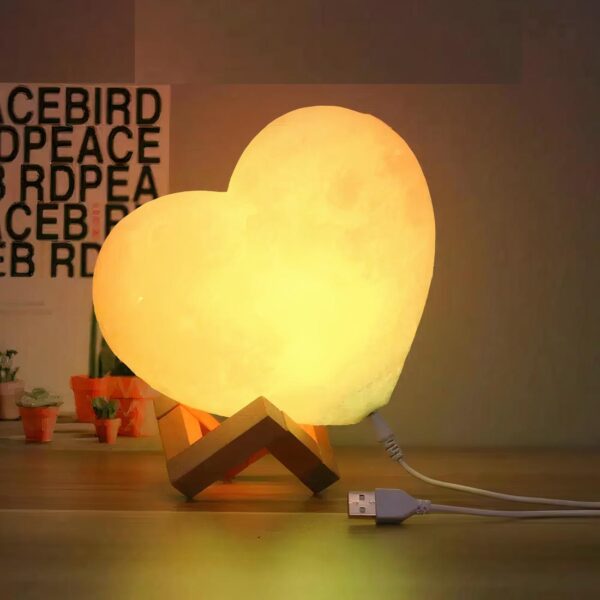 Lampe de chevet fille lune en cœur, allumée et posée sur une table de chevet en bois.
