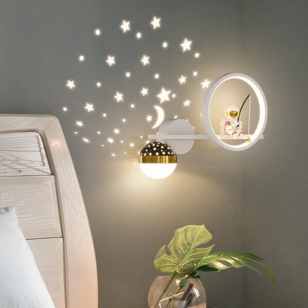 Lampe de chevet murale astronaute accrochée au mur de la chambre d'un enfant et allumée. On voit se refléter les étoiles lumineuses sur le mur gris.