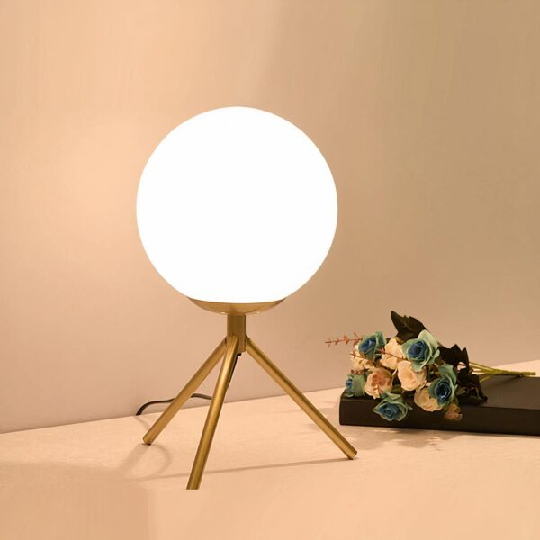 Lampe boule sur trépied en métal, allumée et en version dorée, posée sur un meuble laqué blanc avec des fleurs posées à côté.
