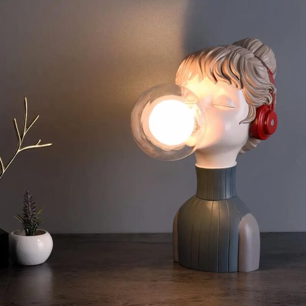 Lampe de chevet buste de jeune fille et bulle d'ampoule, allumée et posée sur un meuble en bois, sur fond gris.