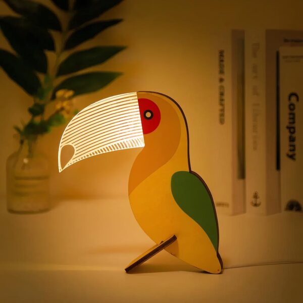 Lampe de chevet colorée en forme de toucan allumée, posée sur un bureau avec des livres en second plan.