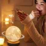 Lampe de chevet Lune rotative allumée devant une femme qui regarde en souriant son téléphone.