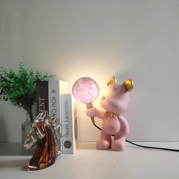 Lampe de chevet ourson kawaii allumée et en version lumière multicolore, sur un meuble laqué blanc, à côté d'une pile de livres et d'une plante.