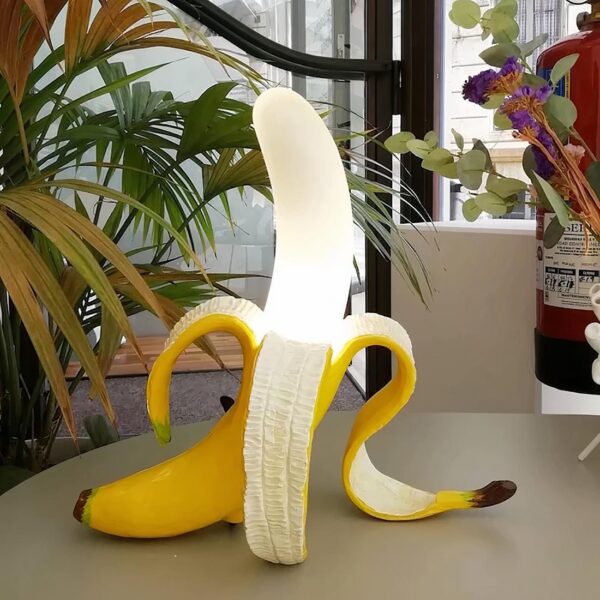 Lampe de chevet Pop-Art banane, allumée et posée sur un meuble au milieu de plusieurs plantes.