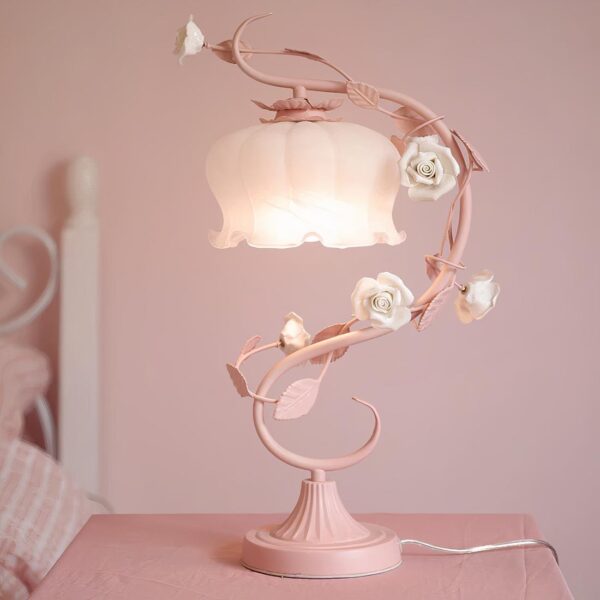 Lampe de chevet romantique en métal et céramique, en version rose, allumée et mise en avant sur une table de chevet, à côté d'un lit, dans une chambre à coucher.