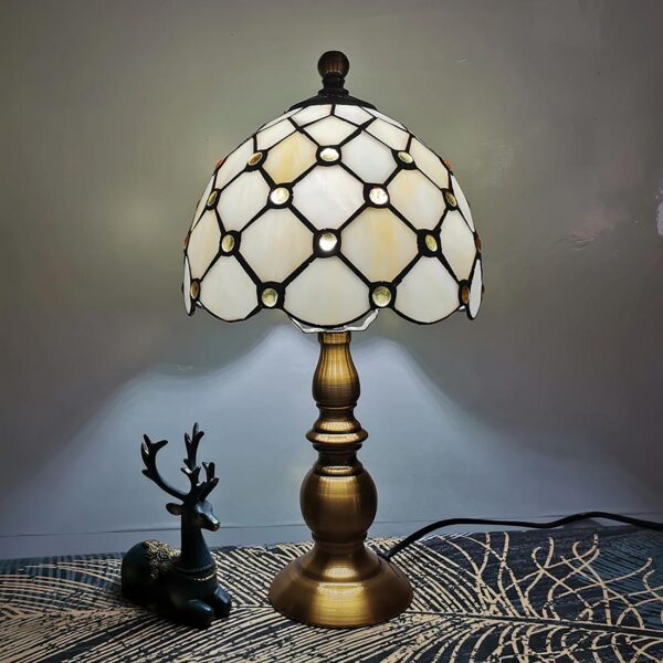 Lampe de chevet style Tiffany au pied doré et abat-jour blanc avec des perles posée sur un support avec un cerf en décoration.