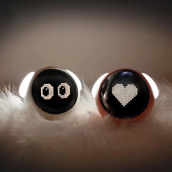 Réveil intelligent lampe de chevet kawaii, allumé, avec l'affichage "yeux", sur un lit de plumes blanches, sur fond gris.