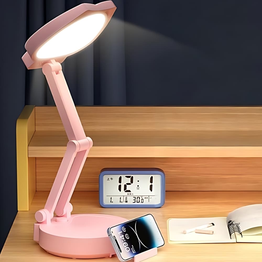 Lampe de chevet fille tactile mignonne avec oreille de chat de couleur rose avec un bras articulé. Posée sur un bureau en bois avec un réveil derrière et un téléphone posé devant. La lumière est allumée.