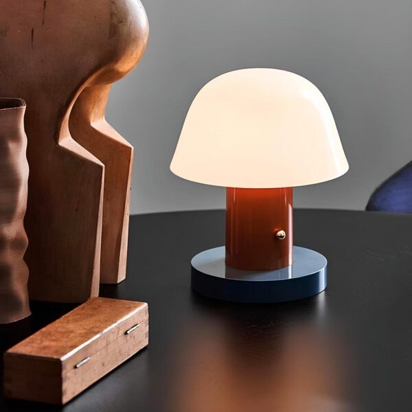 Lampe de chevet sans fil en forme de champignon avec sa base bleu, le pied marron et le chapeau blanc diffusant la lumière. Elle est posée sur un support noir. A gauche se trouvent une boite rectangulaire en bois et une statue en bois.