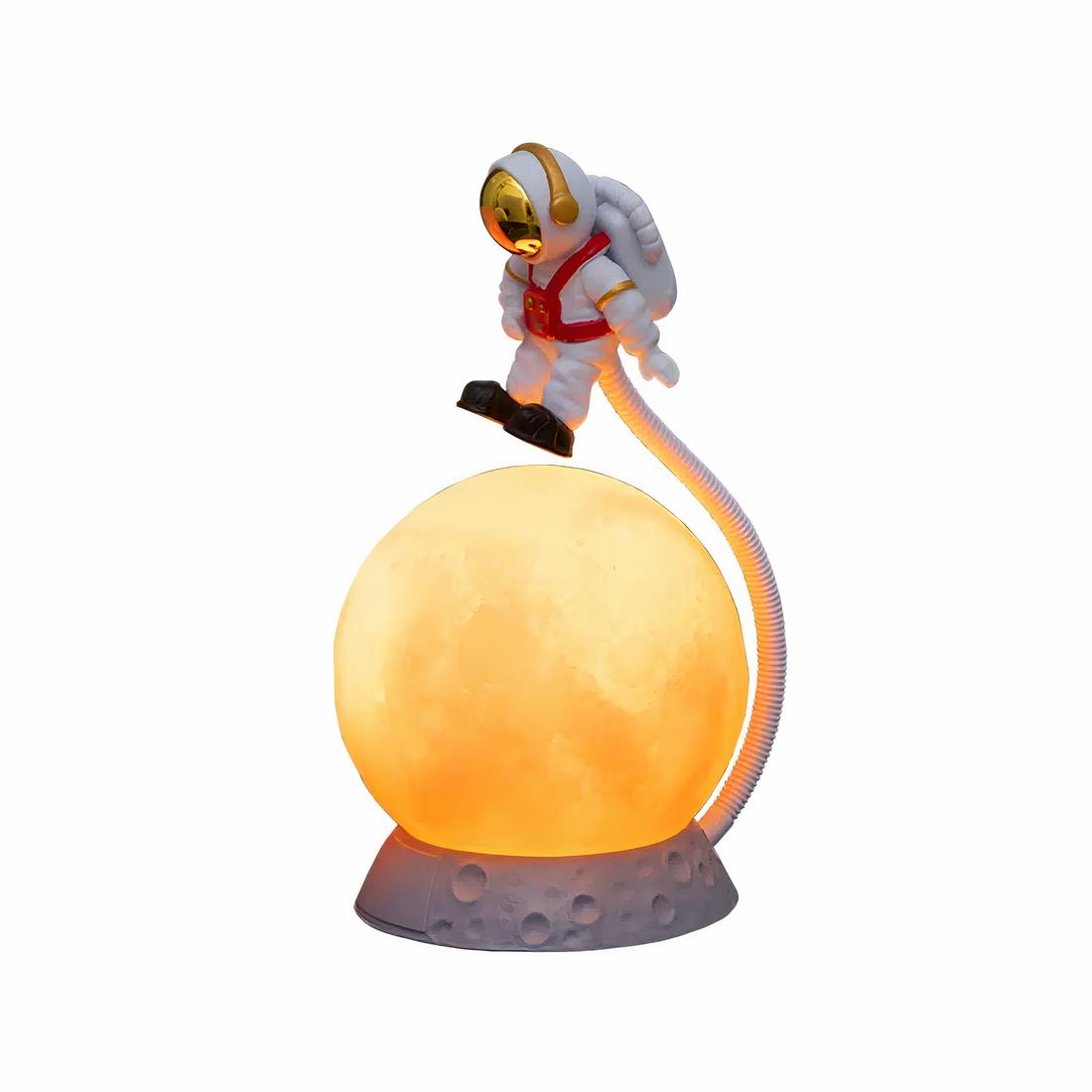Lampe de chevet en forme de lune orange avec un astronaute en apesanteur au-dessus relié à un tuyau blanc à droite. Sur fond blanc.