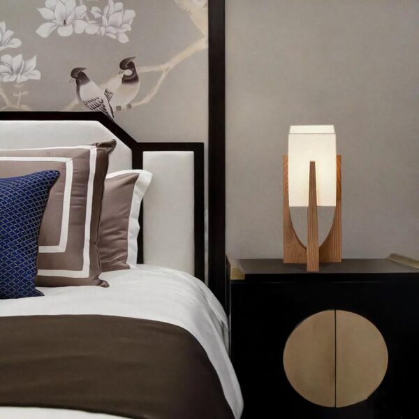 Lampe de chevet avec support verticale en bois tenant en son sommet un rectangle lumineux. Posée sur une table de chevet marron foncé. A gauche, un lit avec tête de lit noire et blanche, literie marron et blanche et un coussin bleu.