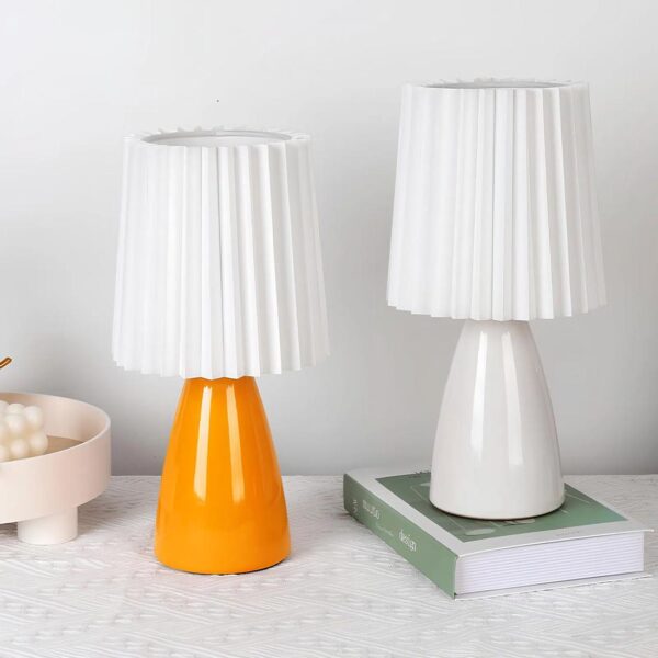 Deux lampes identiques celle de gauche à la pied orange et celle de droite le pied est blanc. Au-dessus, un abat-jour plissé blanc. La lampe blanche est rehaussée sur un livre. blanc et vert. Elles sont sur un tissu blanc.