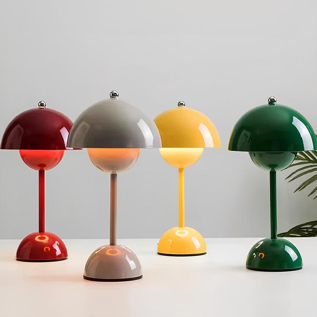 4 lampes de chevet allumées en forme de champignon de couleurs rouge, grise, jaune et verte de gauche à droite. Le socle est en forme de demi boule, relié à la tige fine. Posées sur un support blanc.