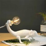 Lampe de chevet en forme de caméléon en résine blanche, qui tient une ampoule dans sa queue, reliée à son câble.Le caméléon est posé sur un document avec des feuillages, ce document est sur un support beige. A droite, un vase transparent avec des feuilles fines et longues.