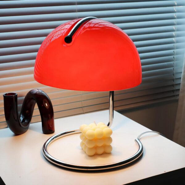 Lampe de chevet moderne de style italien avec structure en métal. Son socle est un cercle ouvert en métal qui remonte pour tenir son abat-jour en verre de couleur rouge. L'ampoule est allumée et éclaire une bougie blanche en cube sous la lampe.Elle est posée sur un support blanc carré, un tube en décoration à gauche et des stores vénitiens derrière.