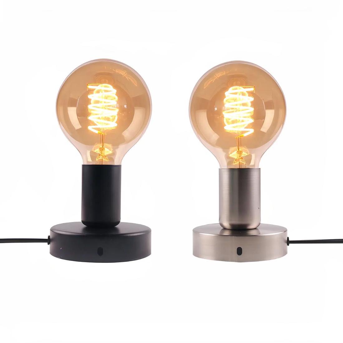 Deux lampes de chevet minimalistes de style industriel. Socle rond, douille et ampoule allumée. Celle de gauche est noire avec son câble vers la gauche, celle de droite est grise avec son câble vers la droite. Sur fond blanc.