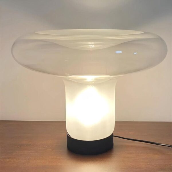 Lampe de chevet en verre en forme de champignon moderne. Le bas du pied est noir, la lumière est allumée et l'ampoule se trouve dans le pied. Elle est posée sur un support couleur bois foncé. Un mur blanc derrière.