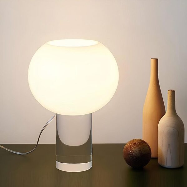 Lampe de chevet moderne. Son socle et un cylindre vertical en verre transparent. Dessus se trouve un ovale blanc avec la lumière diffusée à l'intérieur. La lampe est filaire, son fil se trouve à gauche. Elle est psée sur un support marron. A droite, deux bouteille style en bois et une boule marron. Un mur blanc derrière.
