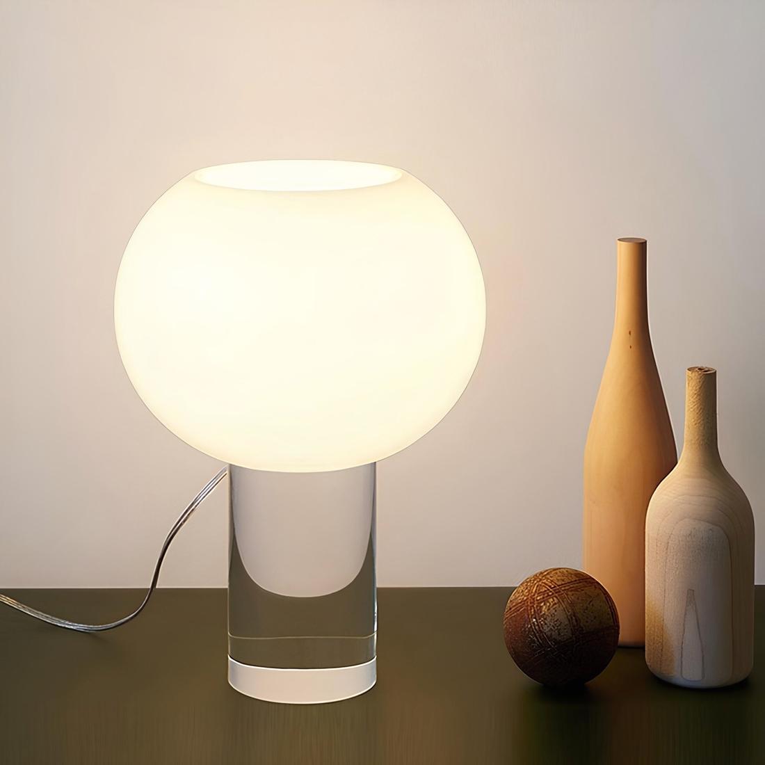 IMG_04_2024_Lampe_chevet_moderne_verre_style_italien_1 Lampe de chevet moderne. Son socle et un cylindre vertical en verre transparent. Dessus se trouve un ovale blanc avec la lumière diffusée à l'intérieur. La lampe est filaire, son fil se trouve à gauche. Elle est psée sur un support marron. A droite, deux bouteille style en bois et une boule marron. Un mur blanc derrière.