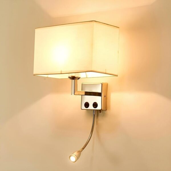 Lampe de chevet murale avec support en métal argenté carré et tige coudée vers le haut . Sur le support il y a 2 interrupteurs. Une lampe de lecture en dessous allumée. Sur la tige coudée, un abat-jour rectangulaire en tissu beige. L'ampoule est allumée. Sur un mur beige.