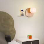 Lampe de chevet murale avec un socle rond couleur bois sur lequel est fixée une lune lumineuse. A gauche, une petit étégère avec un astronaute en équilibre sur une lune grise avec une étoile et une demi-lune dorées. Fixée dans une chambre, au-dessus d'une table de chevet blanche et noire avec un rubik's cube dessus. A gauche, un lit.