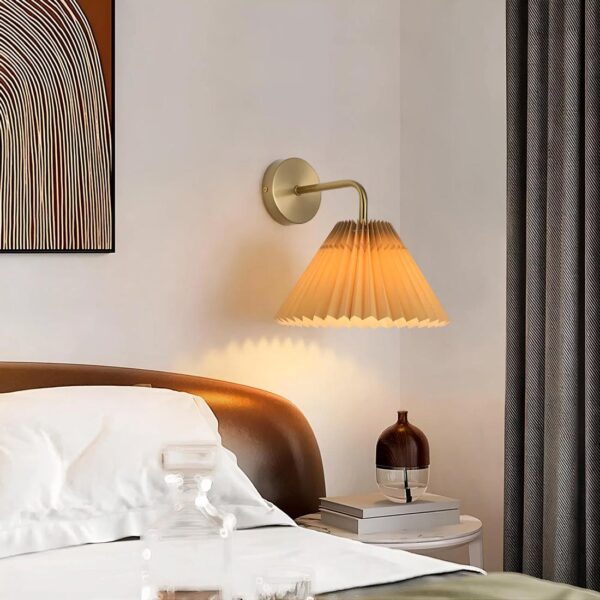 Lampe de chevet murale avec socle rond doré et tige coudée vers le bas avec abat-jours plissé orangé avec ampoule allmuée. Fixée dans une chambre à droite d'un lit avec tête de lit en bois et literie blanche. Au-dessus d'une table de chevet ronde avec des livres empilés. Un rideau gris à droite et un cadre marron à gauche.