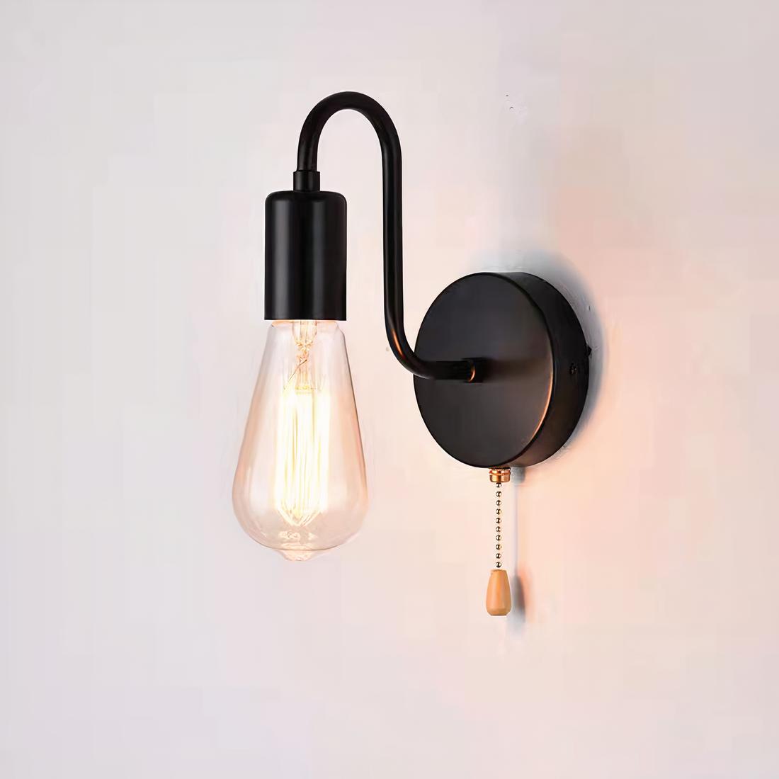 Lampe de chevet murale de style industrielle avec une tige noire en col de cygne. L'ampoule est allumée. Une chaînette se trouve sous le support rond fixé au mur.