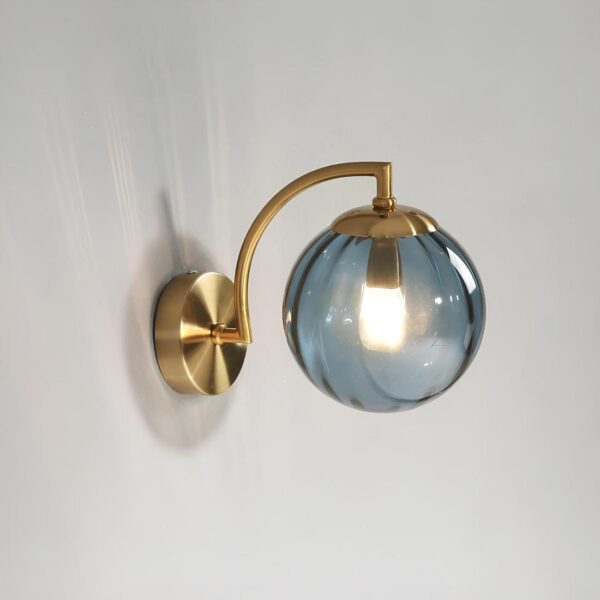 Lampe de chevet murale avec boule en verre bleue laissant voir l'ampoule allumée. Fixée au mur avec un support rond doré et une tige arrondie dorée. Sur un gris.