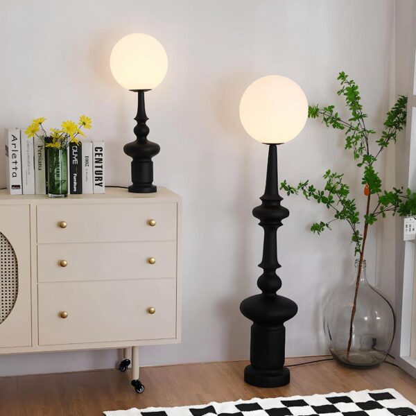 Deux lampes identiques de tailles différentes en forme de lampadaire noir avec boule lumineuse sur le dessus. Un petit modèle se trouve posé sur une commode blanche avec 3 tiroirs sur la gauche. Des livres sont posés à gauche de la lampe. Juste à droite, le même modèle mais plus grand posé au sol à côté de la commode. A sa droite, une boule en verre avec du feuillage. Le sol est de style parquet flottant. Devant la lampe se toruve un tapis style damier noir et blanc.