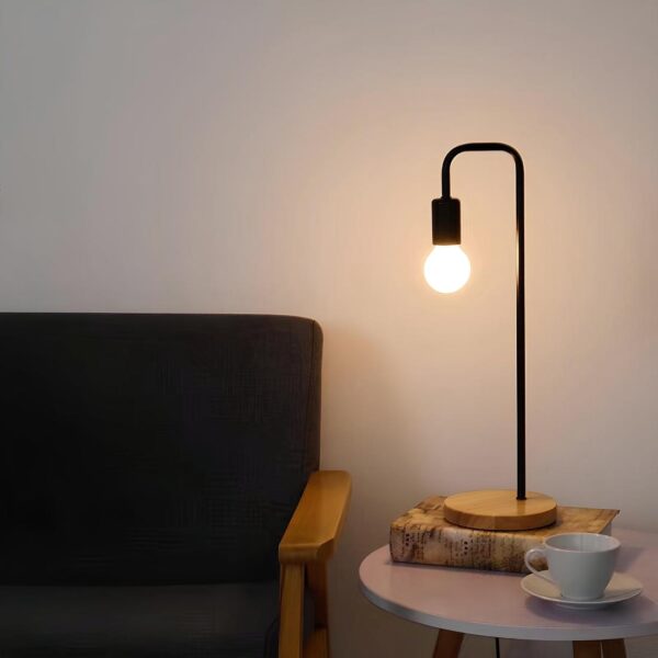 Lampe de chevet de style industriel avec socle rond en bois, tige longue noire coudée et ampoule allumée. Elle est posée sur une table basse rond blanche, sur un livre, à côté une tasse avec sa sous-tasse. A gauche, un canapé noir avec un accoudoir en bois.