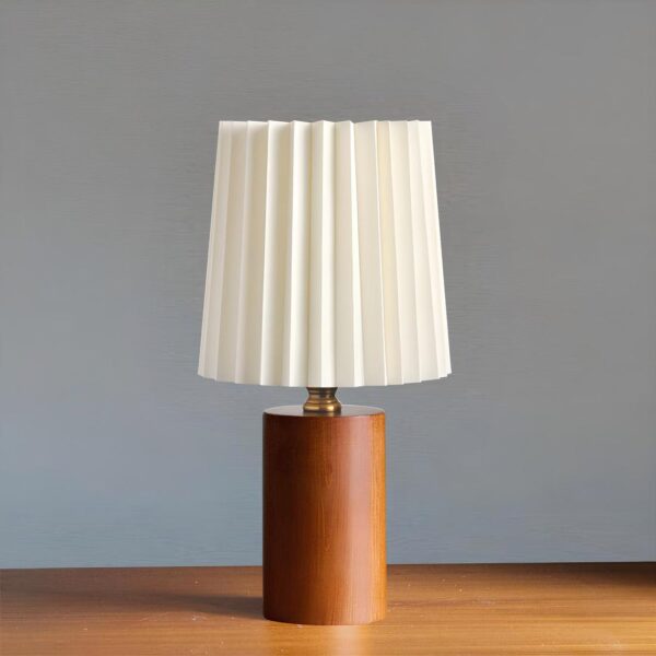 Lampe de chevet avec pied en bois foncé, bague doré en métal au-dessus et abat-jour plissé blanc. Posée sur un support de couleur bois et fond gris.