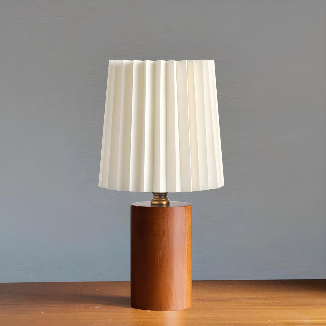 Lampe de chevet avec pied en bois foncé, bague doré en métal au-dessus et abat-jour plissé blanc. Posée sur un support de couleur bois et fond gris.