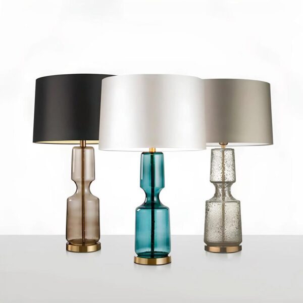 Trois lampes de chevet identiques mais de couleurs différentes. Le pied de la lampe ressemble à un vase en verre translucide, une tige est visible dedans, par-dessus un abat-jour classique. Au fond à gauche, le verre est noir translucide et l'abat-jour noir, Au fond à droite, verre gris translucide et abat-jour gris, devant verre bleu translucide et abat-jour argenté. Les lampes sont sur un socle rond doré. Sur fond blanc et gris pour la partie basse.