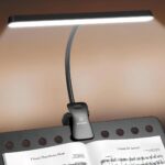 Lampe de lecture noire à pince fixée sur un pupitre noir avec une partition dessus. La lampe est allumée, sa tête d'éclairage est large.
