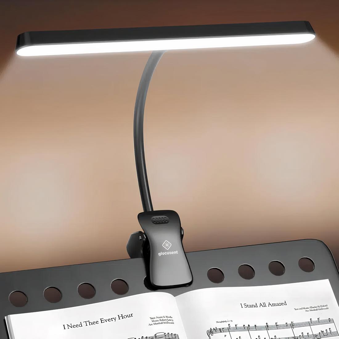 Lampe de lecture noire à pince fixée sur un pupitre noir avec une partition dessus. La lampe est allumée, sa tête d'éclairage est large.
