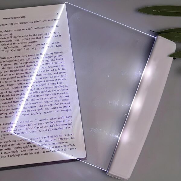 Lampe de lecture plate rectangulaire en plastique transparent. Sa partie de droite est blanche et la lumière est allumée et passe à travers le plastique transparent. Elle est posée à cheval sur une page de livre qui lui-même est posé sur un fond gris.
