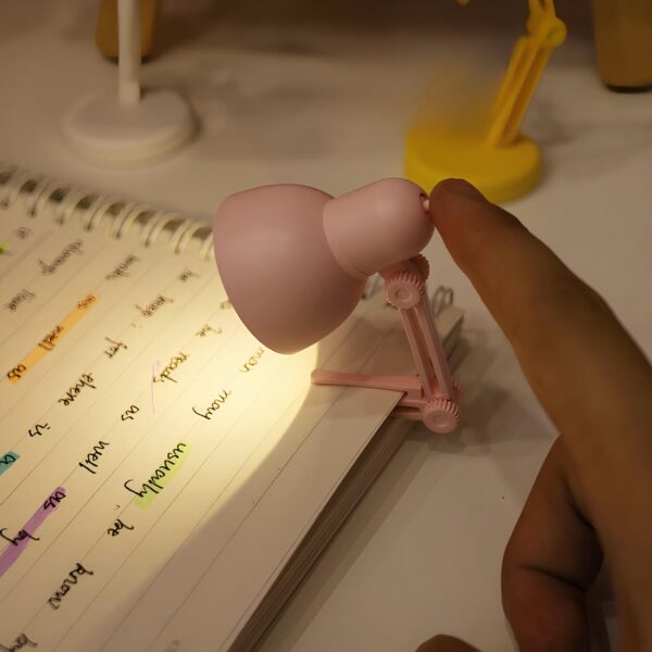Mini lampe de lecture rose pincée sur un cahier et éclairant une page d'écriture. Un doigt touche le bouton de la lampe. En fond on aperçoit une mini lampe blanche et une jaune.