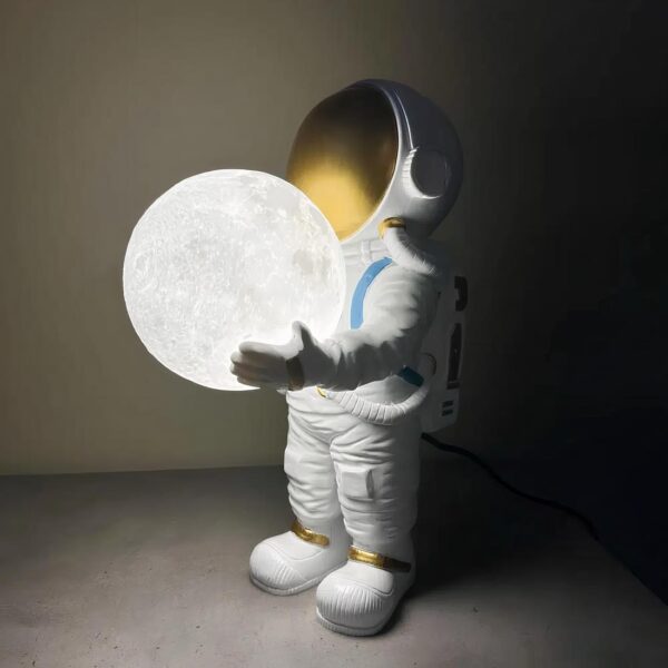 Astronaute en tenue blanche tenant dans ses mains une lune lumineuse. Posé sur un support gris dans la pénombre.
