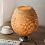 Lampe de chevet avec socle en métal de forme ronde en bambou tressé. Allumée et posée sur une table couleur bois, un document en bas à gauche et une tasse blanche à gauche. Un mur blanc en fond.
