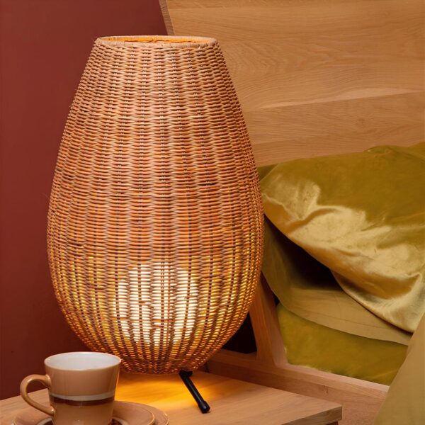Lampe de chevet en rotin en forme d'oeuf avec une boule lumineuse à l'intérieur. Elle est sur pieds fins noirs et posée sur une table de chevet beige avec une tasse à côté. A droite, un lit avec une literie verte anis.