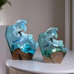 Deux lampes de chevet identiques de 2 tailles différentes : la plus grande à gauche et la petite à droite. Fond sous-marin en résine sur un support en bois. Sous la vague, un requin et une méduse. Sur la vague un surfeur. Lumière allumée passant dans la résine bleue. Les lampes sont posées sur une table ronde blanche.