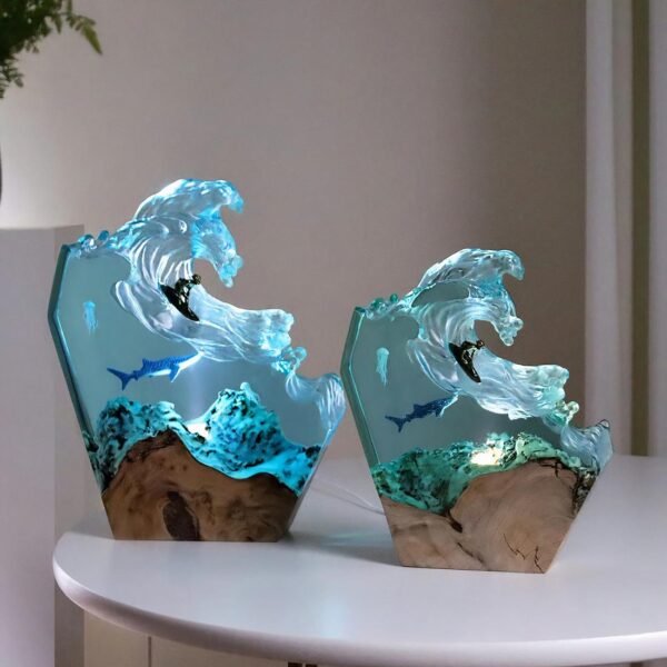 Deux lampes de chevet identiques de 2 tailles différentes : la plus grande à gauche et la petite à droite. Fond sous-marin en résine sur un support en bois. Sous la vague, un requin et une méduse. Sur la vague un surfeur. Lumière allumée passant dans la résine bleue. Les lampes sont posées sur une table ronde blanche.