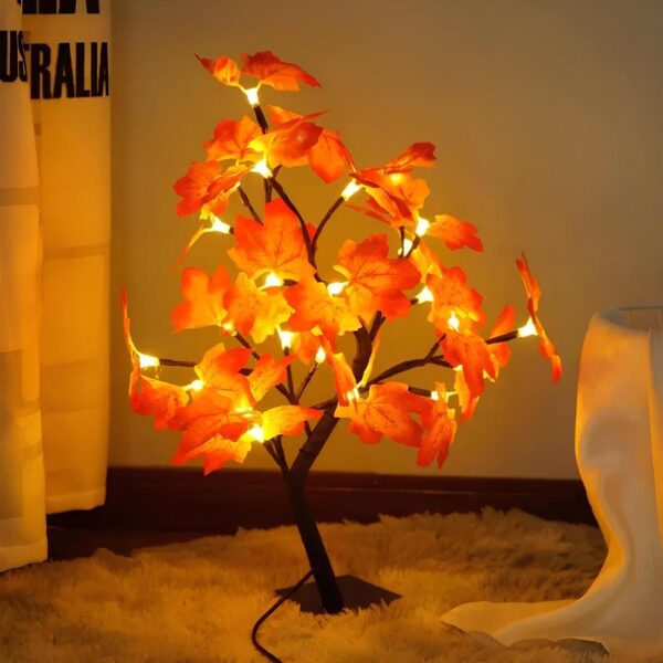 Lampe de chevet filaire en forme d'arbre avec des feuilles d'automne orangées. La lumière se diffuse sous les feuilles pour une lumière orangée dans l'espace. Elle est sur un tapis beige style fourrure, un tissu blanc à droite et un rideau blanc à gauche.