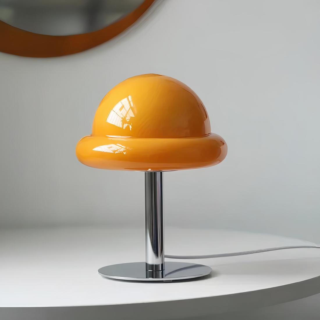 Lampe en forme de champignon, son chapeau est en verre orange, son pied est en métal argenté, relié par un fil. Posée sur un support blanc.