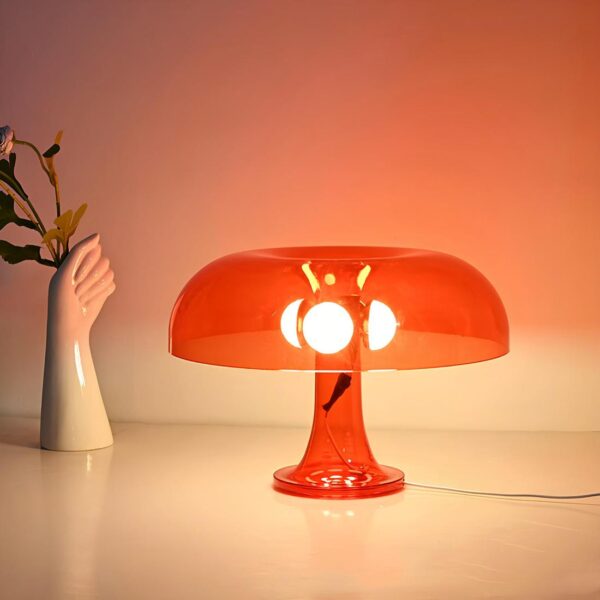 Lampe de chevet en forme de champignon en rouge translucide posée sur un support blanc, elle a 3 sources de lumières sous le chapeau du champignon. A gauche, un vase en forme d'avant-bras avec des fleurs dans la main.