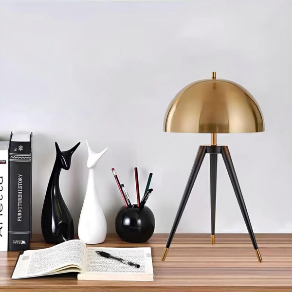 Lampe de chevet en forme de champignon doré sur un trépied noir. Posé sur un bureau couleur bois. A gauche, un cahier ouvert avec un stylo posé dessus. Contre le mur, un pot en crayon noir rond avec 3 crayons, 2 statues de chat et des livres.